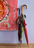 Colourful detail of three walking-stick umbrellas leaning on lavender blue wall next to contemporary painting in hallway