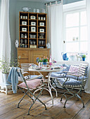 Dining area with white metal table and chairs, patterned cushions and wooden cupboard with shelves and doors