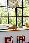 Fischobjekt aus Holz auf moderner Küchentheke und rustikale Barhocker vor Glasfenster mit Sprossen und Gartenblick