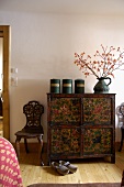 Old cans and pitcher on an antique chest of drawers (painted with flowers), next to a carved, rustic chair