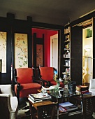 Interior in dark shades with red armchairs and books on table in front of wooden door with Oriental painted motifs