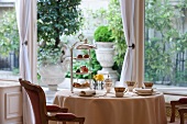 Gedeckter Tisch mit Kaffeegeschirr & Gebäck auf Etagere vor geöffneter Terrassentür mit Blick auf mediterranen Garten