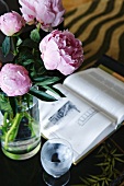 Glass vase with pink peonies next to open book and glass of water