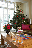Rosenstrauss, Windlichter und Geschenke auf Couchtisch; geschmückter Weihnachtsbaum vor dem Fenster