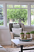 Living room in Scandinavian, country house style; coffee table and comfortable wicker chair below large widow with view of garden