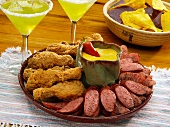 Mexikanische Vorspeisenplatte mit frittierten Hähnchenschenkeln und Wurst (Kielbasa), im Hintergrund Margaritas und Nachos