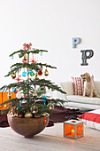 Decorated Christmas tree in bowl and orange glass lantern on wooden floor in front of dog sitting on pale sofa