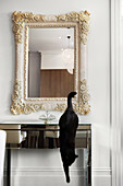Black cat jumping down from mirrored glass console table below wall-mounted mirror with pale wooden frame carved with floral motifs