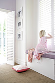 Girl sitting on window seat built into niche of window with closed, white, slatted shutters
