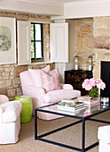 Coffee table with glass top in front of elegant pink armchair with cushions in rustic, modernised living room with stone walls