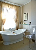 Designer chair and free-standing vintage bathtub below window with curtain in simple bathroom