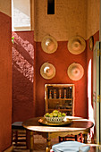 Loggia of Moroccan house with round table in front of straw hats hanging on terracotta wall