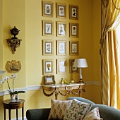 View across modern sofa to antique console table against yellow-painted wall and gilt-framed pictures with floral motifs