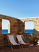 Wooden deckchairs with pale canvas seats on Mediterranean terrace surrounded by high stone wall with openings showing sea view