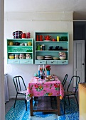 Colourful oilcloth on kitchen table below pastel green shelves on wall