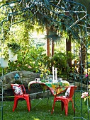 Garden table and red chairs below metal rose arbour with stone fountain in background