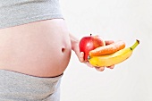 A pregnant woman holding an apple, a banana and a carrot