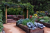 Raised beds with wooden surrounds in lush garden with simple table and benches on wooden decking in background