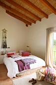 Sichtbare Dachkonstruktion mit Strohmatten in freundlich hellem Schlafzimmer; Kissen und Decken in Rotlilatönen auf französischem Bett