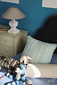 Soft toys, bolster and cushion on bed next to lamp on chest of drawers