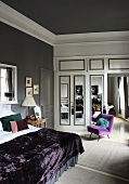 Double bed with purple bedspread, chair with purple upholstery and white fitted wardrobe with mirrored doors in bedroom with grey walls