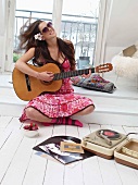 Woman sitting on platform playing the guitar next to retro record player on white floorboards