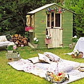 Picknickidylle im Garten mit Patchworkdecke und Vintage-Obstkorb vor blumengeschmücktem Gartenhäuschen