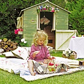 Blonde little girl sitting on patchwork blanket with wire basket of fruit; garden shed decorated with flowers in background