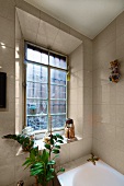 Grünpflanze und kleine Kitschfiguren vor altem Sprossenfenster mit schräger, gefliester Leibung über einer eingebauten Badewanne