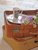 Tasse mit Aufdruck, ein Teller und Blumen auf zwei alten Lederkoffern