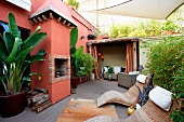 Moderne Rattanliegen und tropische Pflanzen im Kübel auf Terrasse vor mediterranem Wohnhaus mit rot-braun getünchter Fassade