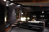 Minimalist dining table and upholstered benches in front of open-plan kitchen in interior in dark tones