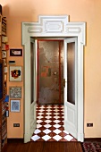 Offene Zimmertür mit geschnitztem weißem Holzrahmen in apricotfarbener Wand und Blick in Vorraum auf Schachbrettmusterboden