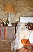 Partially visible sofa with fur blanket next to table lamp on vintage trunk against stone wall