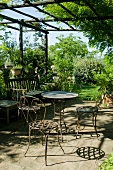Sonnenbeschienene Terrasse und Garten - Tisch und Stühle aus filigranem Metall im Rostlook und Holzsitzbank auf Steinterrasse mit eingewachsener Pergola aus Holz.