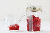 Homemade Strawberry Jam in a Gift Jar with a White Ribbon; Some in a Glass with a Spoon