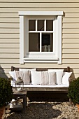 Greyed, weathered teak bench with white cushions below window of wooden house