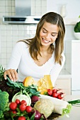 Young woman in kitchen, looking down at assorted fresh vegetables, smiling