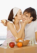 Couple having breakfast on cardboard box, woman kissing man's cheek