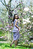 Young woman in dress walking beside flowering tree, looking over shoulder at camera