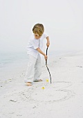 Kleiner Junge am Strand zeichnet mit einem Holzstecken