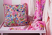 Colourful, ethnic-style cushion on console table next to dresses hanging up