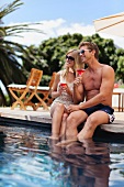 Couple sitting on edge of swimming pool holding drinks