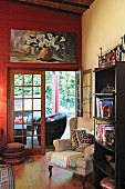 Wing-back chair and bookcase in front of open terrace door; modern still-life painting of calla lilies on red wall
