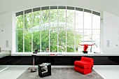 Red armchair and black side table in minimalist interior with huge window