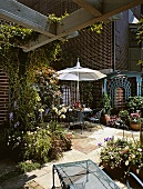 Bewachsene Gartenterrasse eines Backsteinhauses mit vielen Pflanzen, Blumentöpfen, einem Sonnenschirm, rundem Tisch und luftigen Stühlen
