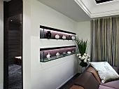 Beleuchtete Wandnischen mit Porzellanvasen auf Glasboarden hinter Sofa im eleganten Wohnraum