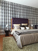Doppelbett mit Bettwäsche in Grautönen und dunkelviolett gepolstertem Kopfteil vor changierendem Vorhang