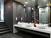 Long mirrored wall above oval washbasins and flower arrangements in Oriental washroom in dark shades