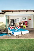Small, Mediterranean bungalow with open glass wall and view into living area beyond; wooden terrace with upholstered loungers in foreground lend a summery holiday feeling